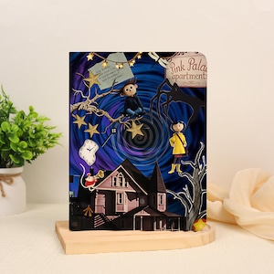 May include: A rectangular notebook featuring a dark blue and purple swirling background with a pink house labeled Pink Palace apartments. It includes illustrations of a character in a yellow raincoat, a character in a dark sweater, a clock, and stars.