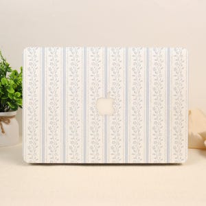 Può includere: Una cover per portatile con un motivo ripetuto di strisce verticali blu e delicati tralci floreali azzurri su sfondo crema, con un ritaglio centrale che rivela il logo Apple argentato sul coperchio del portatile sotto la custodia.