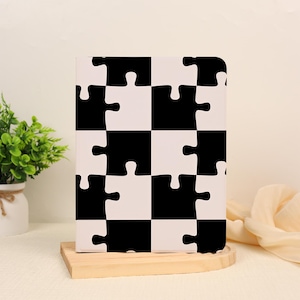 May include: A rectangular notebook featuring a repeating pattern of interlocking black and white puzzle pieces. The cover displays a high-contrast grid design where each square segment is shaped like a jigsaw piece, creating a seamless visual.