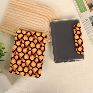 May include: A rectangular ink pad case featuring a repeating pattern of orange and yellow flame characters with smiling faces on a black background. The case has a clear plastic lid and a matching patterned flap that folds over the side of the container.