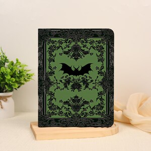 May include: A rectangular notebook with a sage green cover featuring a black bat silhouette in the center. The cover is decorated with a black ornate floral border pattern that frames the central bat graphic on all four sides of the notebook.