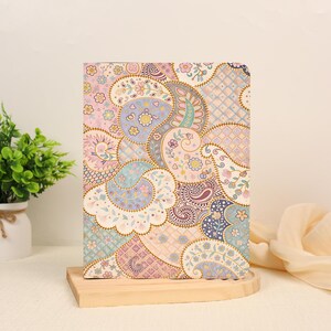 May include: A rectangular notebook featuring a colorful paisley and floral pattern in shades of pink, blue, yellow, and purple. The design includes intricate swirls, small blossoms, and a grid-like texture, all printed on the front cover of the item.