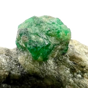 May include: A close-up of a rough-cut emerald gemstone. The emerald is a vibrant green color with a slightly textured surface. It is set against a backdrop of a gray and white rock.