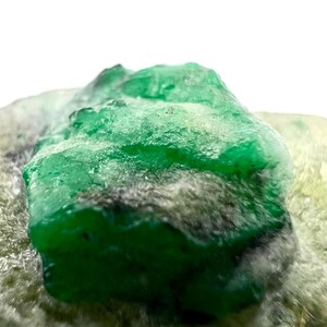 May include: A close-up of a raw emerald gemstone, displaying a rich, deep green colour. The uncut stone is set against a backdrop of a lighter, textured mineral, highlighting its natural beauty and crystalline structure.