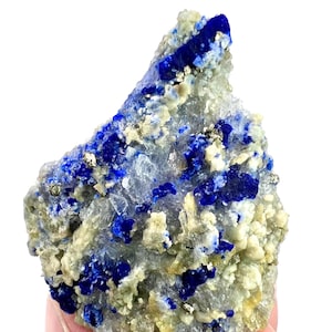 May include: A close-up of a natural lapis lazuli specimen. The rock features deep blue patches and veins, interspersed with flecks of gold-colored pyrite and a matrix of light gray and beige minerals. The overall texture is rough and crystalline.