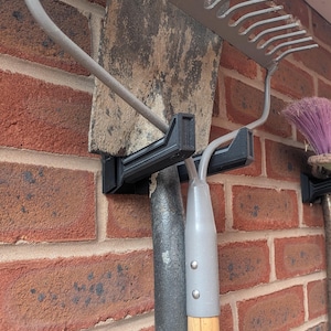 May include: A black plastic wall-mounted storage bracket holding a metal shovel and a metal rake. The bracket features two distinct slots designed to secure the handles and heads of garden tools against a red brick wall surface for organization.