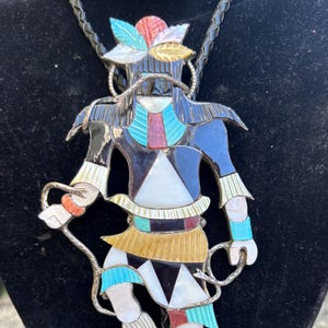 May include: A bolo tie featuring a large, intricate inlay pendant depicting a figure in motion. The pendant is crafted with multicolored stone and shell segments, including turquoise, red, black, and white, set in silver, suspended on a black cord.