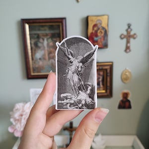 May include: A die-cut sticker featuring a black and white detailed engraving illustration of an angel with large wings standing over a fallen figure while holding a long spear, set against a dark circular background within an arched frame.