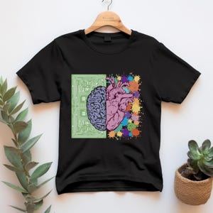 May include: Black t-shirt featuring a graphic design of a brain and a heart. The brain is depicted in purple, set against a green circuit board background. The heart is pink, with colorful paint splatters.