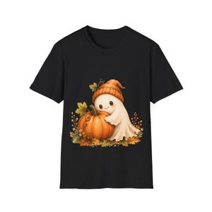 Puede incluir: Una camiseta negra de manga corta con un estampado gráfico de un pequeño fantasma blanco que lleva un gorro de punto naranja. El fantasma abraza una gran calabaza naranja rodeada de hojas de otoño y pequeñas flores en tonos marrones.