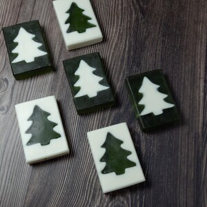 Op de afbeelding: Rechthoekige zeepblokken met een kerstboomontwerp. De zepen wisselen af tussen een witte basis met een groene boom en een groene basis met een witte boom. Ze zijn gerangschikt op een donker houten oppervlak.