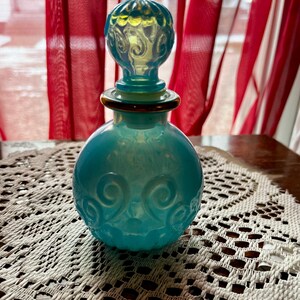 May include: A turquoise glass perfume bottle with a decorative stopper. The bottle has a round body with embossed swirls and a gold-colored band around the neck. The stopper is a lighter shade of turquoise with a swirl design.