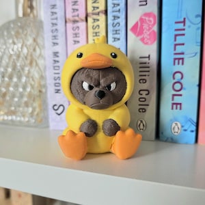 Puede incluir: Una pequeña figura impresa en 3D de un oso marrón con cara de mal humor, vestido con un disfraz de pato amarillo brillante con pico naranja y patas palmeadas naranjas. El oso tiene el ceño fruncido y una expresión severa, sentado erguido.