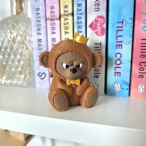 Puede incluir: Una pequeña figura de oso marrón que lleva una corona amarilla y una pajarita amarilla a juego. El oso tiene una superficie texturizada, orejas redondeadas y una expresión facial gruñona con ojos blancos y una nariz negra.