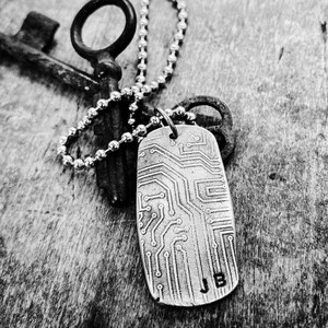 May include: A silver chain necklace with a silver key charm and a silver dog tag pendant with a circuit board design and the initials "JB" engraved on it.