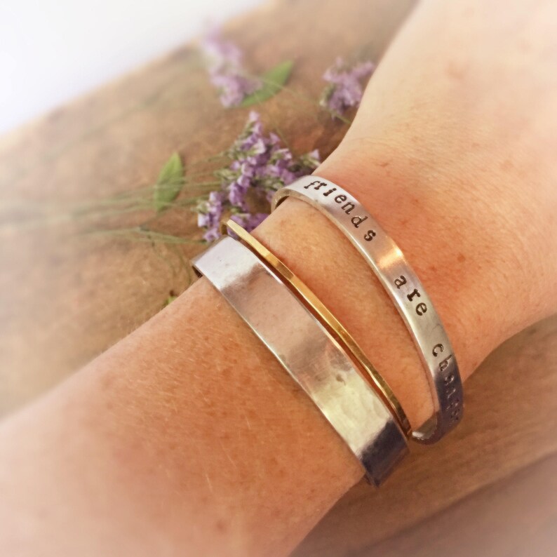 May include: Two silver bracelets on a wrist. The top bracelet is engraved with the words "Friends are chosen".