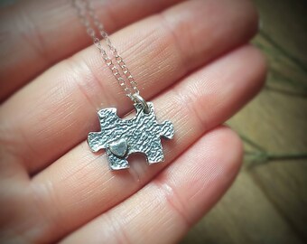 Puede incluir: Un collar de plata con un colgante de pieza de rompecabezas con un pequeño corazón. El colgante es texturizado y tiene un aspecto rústico.