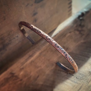 May include: A copper-colored cuff bracelet with an intricate, repeating pattern of swirling lines. The open-ended bracelet is displayed on a wooden surface, showcasing its polished finish and detailed design. The bracelet's warm tones and elegant pattern suggest a handcrafted aesthetic.