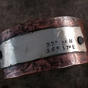 Puede incluir: Una pulsera de cobre y plata con textura martillada. La banda de plata tiene coordenadas grabadas: 37° 31' N 150° 17' E.