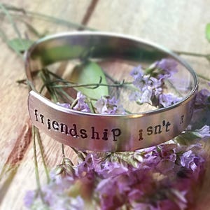 May include: A silver bracelet with the inscription "friendship isn't a" on a wooden surface with purple flowers.
