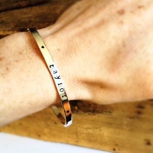 Może przedstawiać: Srebrna bransoletka typu bangle z wygrawerowanym imieniem "taylor". Bransoletka ma mały wisiorek w kształcie serca.