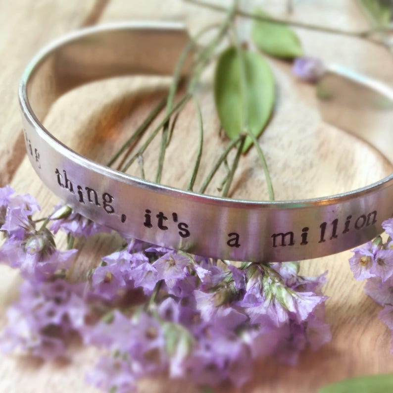 May include: Silver bangle bracelet with the words "little thing, it's a million" engraved on it. The bracelet is lying on a bed of purple flowers.