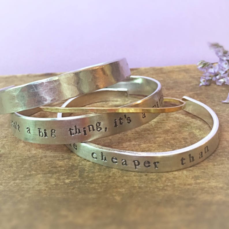 May include: Four silver bracelets stacked on top of each other. The top bracelet has a hammered finish. The bottom bracelet has the text "It's a big thing, it's cheaper than" engraved on it.