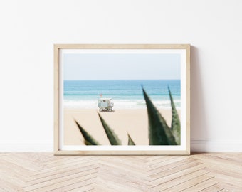 May include: A framed photograph of a lifeguard stand on a sandy beach with blue ocean waves in the background. The stand is white with a red flag on top. The photo is framed in a light wood frame and is partially obscured by a green plant in the foreground.