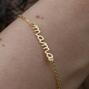 May include: A delicate gold bracelet featuring the word "mamas" in cursive lettering. The fine chain bracelet is worn on a wrist. The bracelet is a simple, elegant piece.