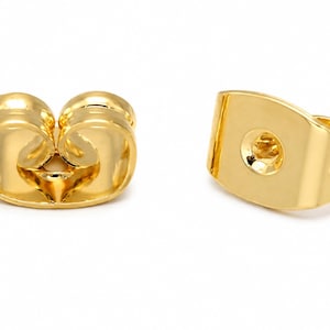 May include: Two gold-colored earring backs with a polished finish. One piece is shown from the side, displaying a curved, rounded shape, while the other is positioned to reveal its flat, rectangular face with a central circular indentation.