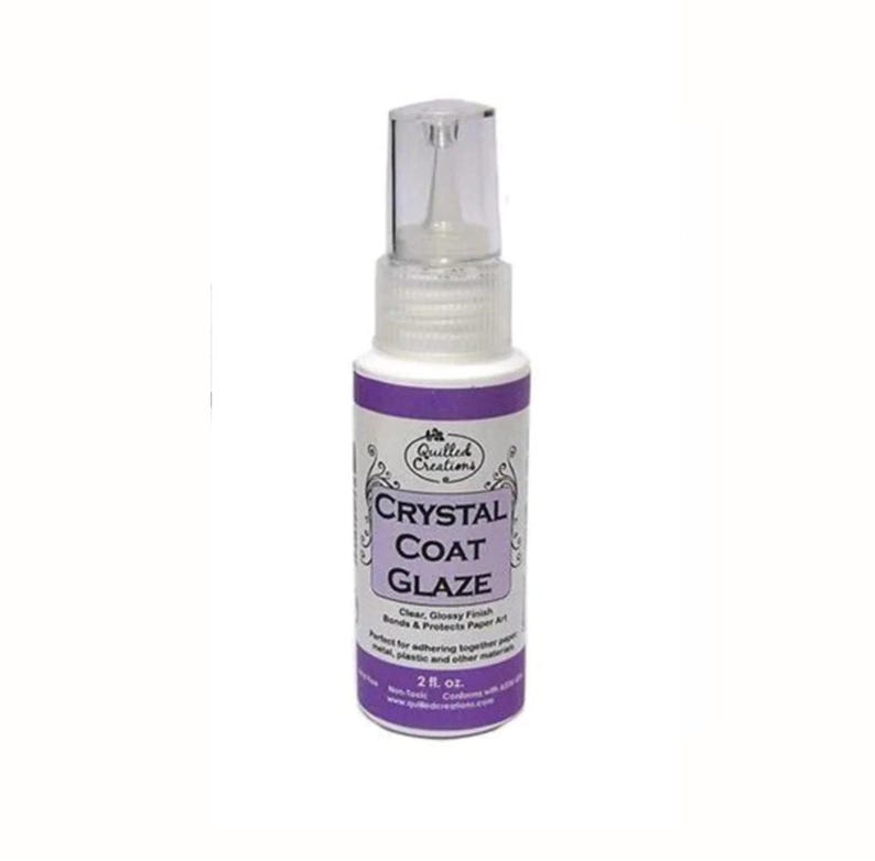 Op de afbeelding: Een witte plastic spuitfles van Quilled Creations Crystal Coat Glaze met een doorzichtige dop. Het etiket is paars en wit met de tekst Crystal Coat Glaze, Clear, Glossy Finish, Bonds & Protects Paper Art en 59 ml erop gedrukt.