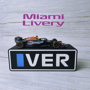 May include: A black and red Formula 1 race car model with sponsor logos, displayed on a black base with the word "VER" in white. The text "Miami Livery" is above the car.