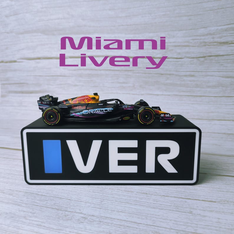 May include: A black and red Formula 1 race car model with sponsor logos, displayed on a black base with the word "VER" in white. The text "Miami Livery" is above the car.