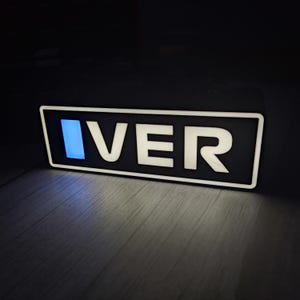 May include: A rectangular sign with the word "IVER" in white, illuminated letters against a black background. The letter "I" has a blue glow. The sign is on a wooden surface.