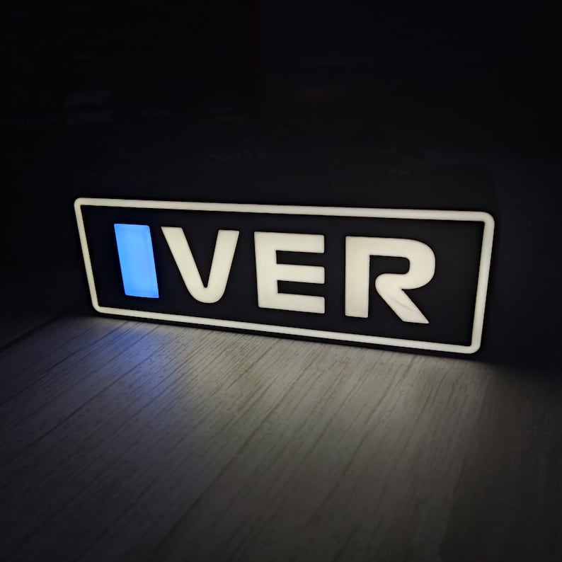 May include: A rectangular sign with the word "IVER" in white, illuminated letters against a black background. The letter "I" has a blue glow. The sign is on a wooden surface.