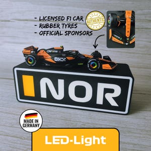 Könnte beinhalten: Ein schwarzes und orangefarbenes F1-Rennwagenmodell mit Gummireifen, präsentiert auf einer schwarzen LED-Lichtbasis. Die Basis trägt das Wort "NOR" in Weiß und einen orangefarbenen Streifen. Der Text lautet "Lizenzierter F1-Wagen" und "Offizielle Sponsoren".