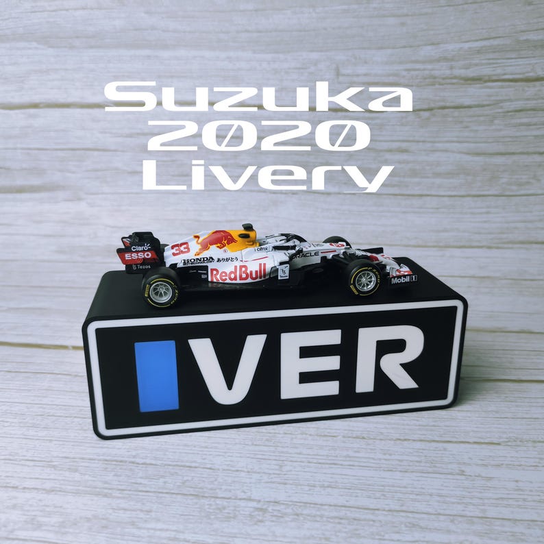 May include: A miniature Red Bull racing car model, white with red and yellow accents, sits on a black display stand. The stand has the word "VER" in white and a blue square. The text "Suzuka 2020 Livery" is above the car.