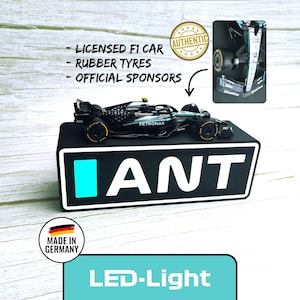 Peut inclure: Une maquette de voiture de course F1 noire et sarcelle avec des pneus en caoutchouc, exposée sur une base noire avec le mot "ANT" en blanc. L'image comprend le texte "Licensed F1 Car", "Rubber Tyres" et "Official Sponsors".