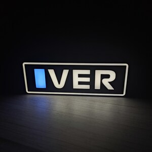 May include: A rectangular sign with the word "IVER" in white, illuminated letters. The letters are set against a black background within a white border. A blue vertical rectangle is on the left side of the sign.
