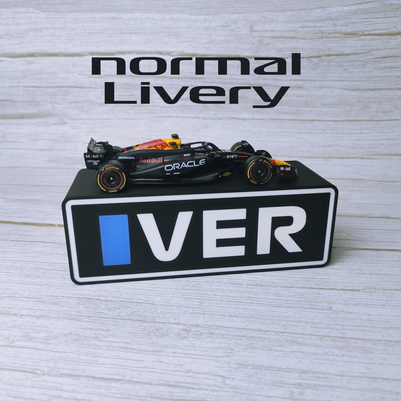 May include: A black and red Formula 1 race car model with sponsor logos, displayed on a black stand with the word "VER" in white. The text "normal Livery" is above the car. The car has yellow and red accents.