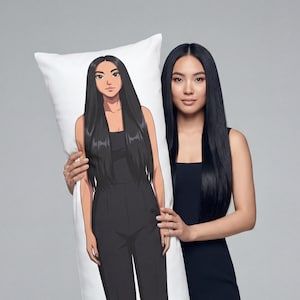 May include: A white body pillow featuring a full-body illustration of a person in a black jumpsuit. The pillow is held by a person wearing a black jumpsuit. The background is a solid light gray.