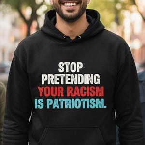 以下が含まれることがあります： 白、赤、青で「STOP PRETENDING YOUR RACISM IS PATRIOTISM」と書かれた黒いフード付きスウェットシャツ。 画像には、並木道を歩く3人の人々が写っています。