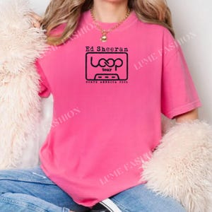 May include: A bright pink t-shirt featuring the text "Ed Sheeran Loop tour North America 2026" in a black graphic. The design is a stylized cassette tape. The shirt is a short-sleeved, casual style.