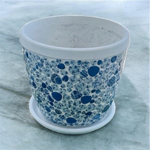 Puede incluir: Una maceta de cerámica con borde y platillo blancos. La maceta está decorada con un patrón de mosaico de baldosas circulares azules y blancas. Las baldosas tienen diseños florales y abstractos.