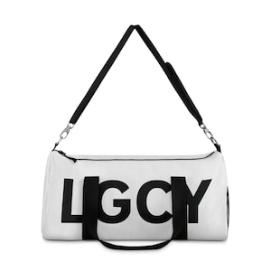 Puede incluir: Bolsa de lona blanca con asas negras y una correa para el hombro negra. La bolsa presenta la palabra "LGCY" en letras mayúsculas negras grandes y en negrita en la parte delantera. La bolsa tiene forma cilíndrica y está diseñada para transportar equipos deportivos o artículos de viaje.