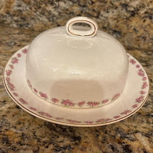 May include: A white ceramic butter dish with a domed lid and a matching plate. The rim of the plate and the lid are decorated with a pink floral pattern and a gold trim. The lid has a small handle.