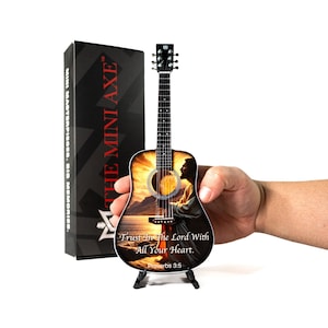 May include: A miniature guitar with a religious landscape design, held in a person's hand. The guitar features the text "Trust In The Lord With All Your Heart. Proverbs 3:5." A black box with "THE MINI AXE" is in the background.