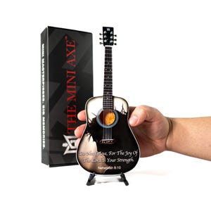 May include: A miniature acoustic guitar with a black and white design, held in a hand. The guitar features a landscape scene and the text "Do Not Grieve, For The Joy Of The Lord Is Your Strength. Nehemiah 8:10". A black box with the text "THE MINI AXE" is in the background.