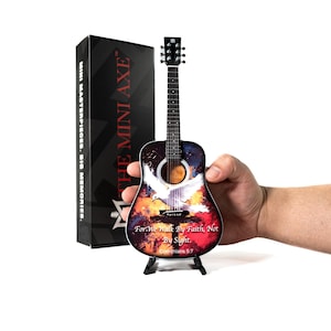 May include: A miniature acoustic guitar with a colorful design featuring a dove and text from Corinthians 5:7. The guitar is displayed on a small black stand, with a black box labeled "THE MINI AXE" in the background.