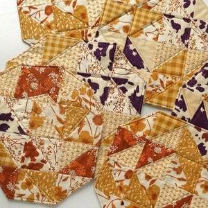 May include: A collection of handmade, geometric fabric coasters. The coasters feature a patchwork design with various patterns, including floral, gingham, and cow print, in shades of orange, yellow, and purple. The coasters are arranged on a white surface.
